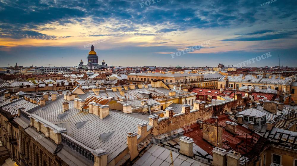 Фотография показывает великолепный вид на крыши Санкт-Петербурга с возвышающимся вдали куполом Исаакиевского собора. Рассеянные облака в небе и мягкие оттенки заката или восхода придают изображению поэтическую атмосферу. Кровли зданий имеют различные оттенки и текстуры, что создаёт уникальный городской пейзаж. Такой снимок может быть использован для иллюстрации книг и статей о Санкт-Петербурге, в арт-проектах, а также как элемент декора.